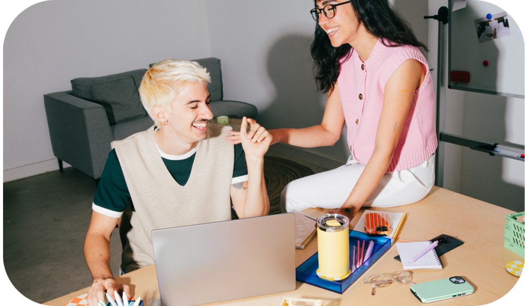 Ki Jugendhilfe Förderprogramm jetzt anmelden - Junge Menschen sind am Schreibtisch und lachen beide ausgelassen. Auf dem Schreibtisch bedfinden sich unterschiedliche Gegenstände, wie Latop, Becher und Stifte.